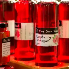Flaschen mit rotem Himbeeressig von "The Jam Shed". Ein Glas mit ähnlichem Inhalt steht davor. Was ist Tafelessig? Hier ist ein Beispiel für Fruchtessig.