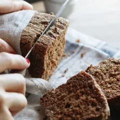 Hände schneiden ein dunkles, gesundes Roggenbrot. Mehrere Scheiben liegen auf einem Leinentuch.