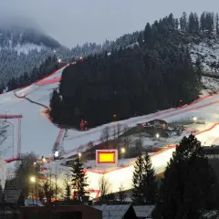 Die Skirennstrecke, beleuchtet für die Nacht, mit roten Banden und einem roten Hahnkamm-Symbol.