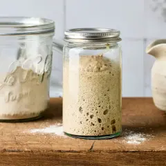 Ein Glas mit aktivem Sauerteig, bereit zum Anstellgut auffrischen. Daneben ein Glas mit Mehl und eine Kanne.