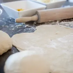 Pizzamehl liegt auf dem Tisch, Teigkugeln und ausgerollter Teig warten auf Belag.