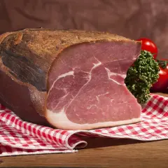 Ein Stück Katenschinken, rustikal gereift, mit saftigem Fleisch und weißem Fett. Daneben Kirschtomaten und Petersilie auf kariertem Tuch.