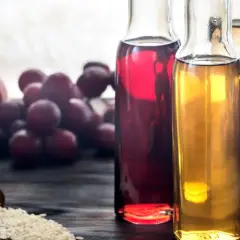 Flaschen mit verschiedenen Essigsorten, darunter dunkler Balsamico-Essig, daneben Äpfel, Trauben und eine Schaufel Reis.