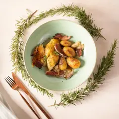 Ein Teller mit Hähnchen, Kartoffeln und Speck, umrahmt von Rosmarinzweigen. Die Rosmarin verwendung verleiht dem Gericht ein wunderbares Aroma.