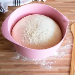 Ein aufgegangener Hefeteig mit Milch in einer rosa Schüssel, bereit zum Backen. Daneben liegt ein Holzlöffel mit Herz.