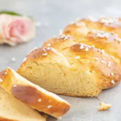 Ein goldbraunes Zopf Brot, bestreut mit Hagelzucker, liegt auf einem Tisch. Daneben eine rosa Rose und eine Tasse.
