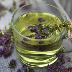 Ein Glas mit goldenem Thymiantee, umgeben von frischen Thymianblüten. Vielleicht ein bisschen zu viel Thymiantee für einen Schluck.