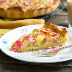 Ein Stück Rhabarber Rahm Kuchen auf einem Teller mit Blumenmuster, daneben ein Glas Kaffee.