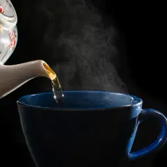Heißer Estragon Tee wird aus einer blumengeschmückten Kanne in eine blaue Tasse gegossen. Dampf steigt auf.