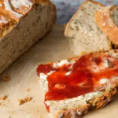 Frisch aufgebackenes Brot mit Butter und Marmelade auf einem Holzbrett. Ein Messer liegt daneben.