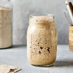 Blubbernder Sauerteig im Glas, bereit zum Backen. Daneben ein Glas mit Mehl und Küchenutensilien.