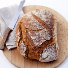 Ein frisch gebackenes Brot aus Hefe Teig liegt auf einem Holzbrett. Daneben ein Messer und eine Serviette.