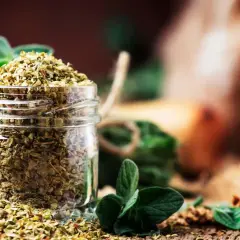 Ein Glas voll getrocknetem Oregano, bereit für die Küche. Frische Zweige im Hintergrund erinnern an das Aroma, das beim Oregano trocknen entsteht.