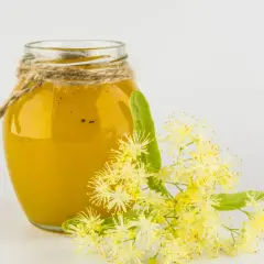 Ein Glas goldgelber Lindenhonig, verziert mit Juteschnur, neben frischen Lindenblüten. Lindenhonig gesund und wohltuend.
