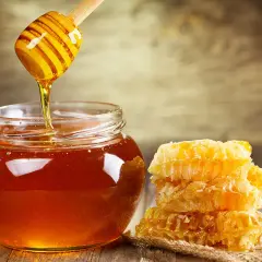 Goldener Honig tropft von einem Holzstäbchen in ein Glas. Daneben ein Stapel Waben. Die Frage, wie lange ist honig haltbar, wird hier visuell beantwortet: ewig!