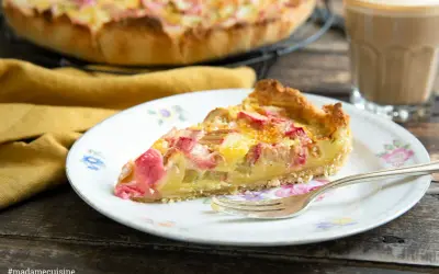Ein Stück Rhabarber Rahm Kuchen auf einem Teller mit Blumenmuster, daneben ein Glas Kaffee.