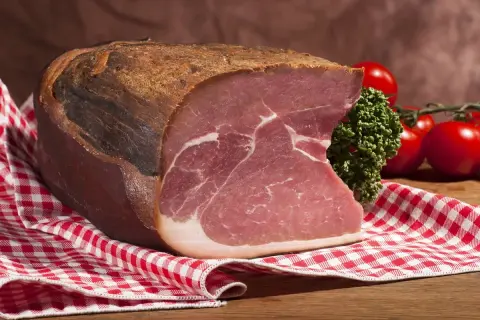 Ein Stück Katenschinken, rustikal gereift, mit saftigem Fleisch und weißem Fett. Daneben Kirschtomaten und Petersilie auf kariertem Tuch.