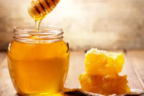 Ein Honigheber tropft goldenen Manuka Honig in ein Glas. Daneben liegt ein Stück Wabe. So schmeckt manuka honig!
