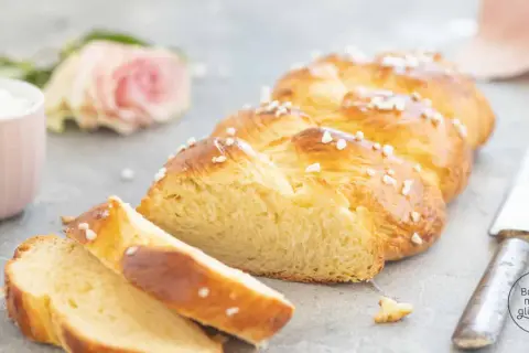 Ein goldbraunes Zopf Brot, bestreut mit Hagelzucker, liegt auf einem Tisch. Daneben eine rosa Rose und eine Tasse.