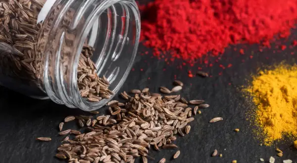 Braune Kümmelsamen quellen aus einem Glas, daneben leuchtend rote und gelbe Gewürzpulver. Was ist Cumin? Es ist dieses aromatische Gewürz!