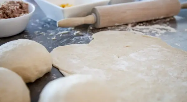 Pizzamehl liegt auf dem Tisch, Teigkugeln und ausgerollter Teig warten auf Belag.