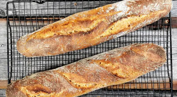 Zwei knusprige, goldbraune Sauerteig Baguettes liegen auf einem schwarzen Gitterrost.