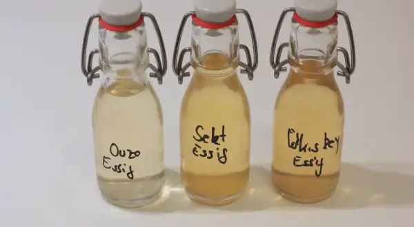 Drei kleine Flaschen mit Bügelverschluss, gefüllt mit Flüssigkeiten. Beschriftet sind sie mit "Ouzo Essig", "Sekt Essig" und "Whiskey Essig".