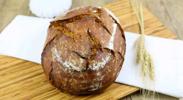 Ein rustikales Bierbrot mit Sauerteig, auf einem Holzbrett mit einem weißen Tuch und Ähren dekoriert.