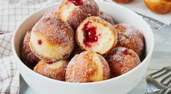 Schüssel voller süßer Berliner, bestreut mit Zucker. Ein Berliner ist aufgeschnitten und zeigt die rote Marmeladenfüllung. Perfekt für den Karneval!