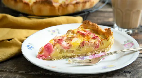 Ein Stück Rhabarber Rahm Kuchen auf einem Teller mit Blumenmuster, daneben ein Glas Kaffee.