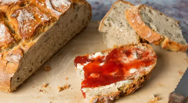 Frisch aufgebackenes Brot mit Butter und Marmelade auf einem Holzbrett. Ein Messer liegt daneben.