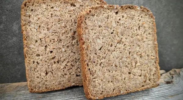 Zwei Scheiben rustikales **Kornbrot** liegen auf Holz. Das Brot ist dicht und voller Körner, mit einer dunklen Kruste.