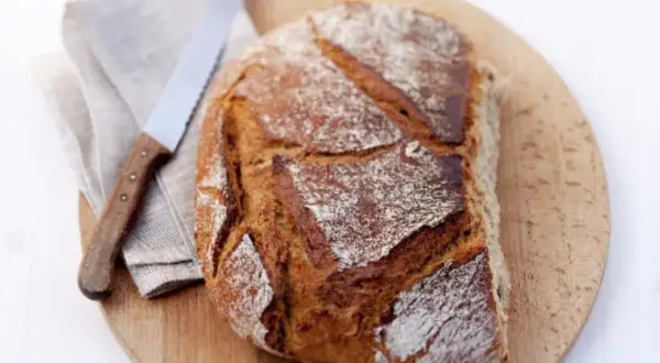 Ein frisch gebackenes Brot aus Hefe Teig liegt auf einem Holzbrett. Daneben ein Messer und eine Serviette.