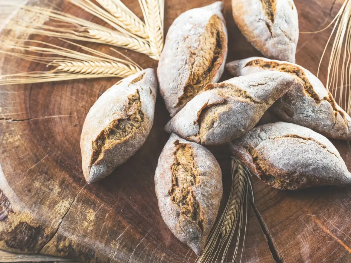 Frische Brötchen, gebacken aus verschiedenen Getreidearten, liegen auf einem rustikalen Holzbrett, umgeben von Ähren.