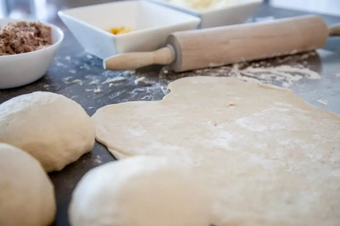 Pizzamehl liegt auf dem Tisch, Teigkugeln und ausgerollter Teig warten auf Belag.