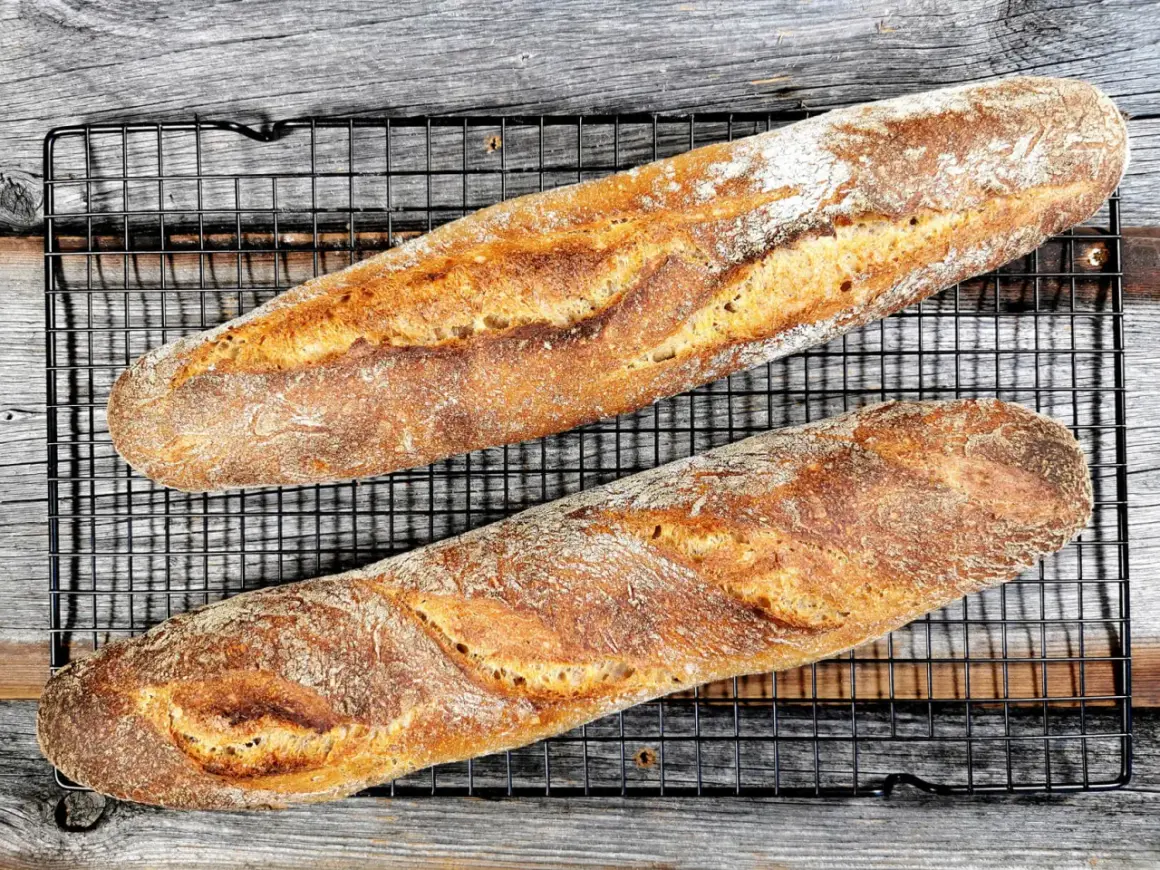 Zwei knusprige, goldbraune Sauerteig Baguettes liegen auf einem schwarzen Gitterrost.