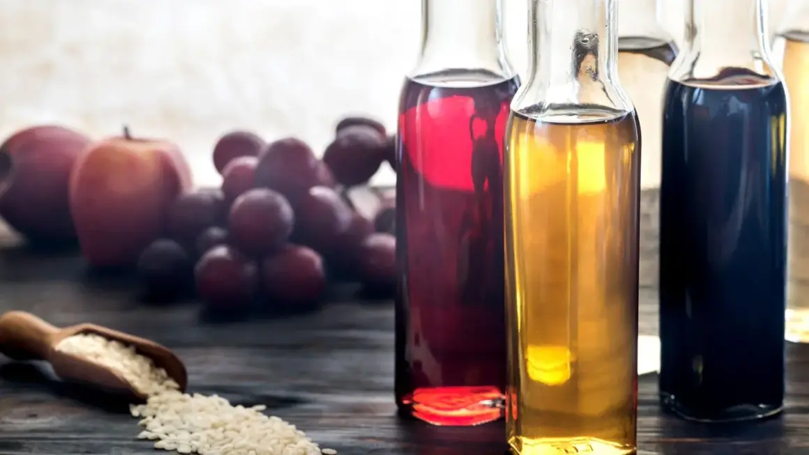 Flaschen mit verschiedenen Essigsorten, darunter dunkler Balsamico-Essig, daneben Äpfel, Trauben und eine Schaufel Reis.