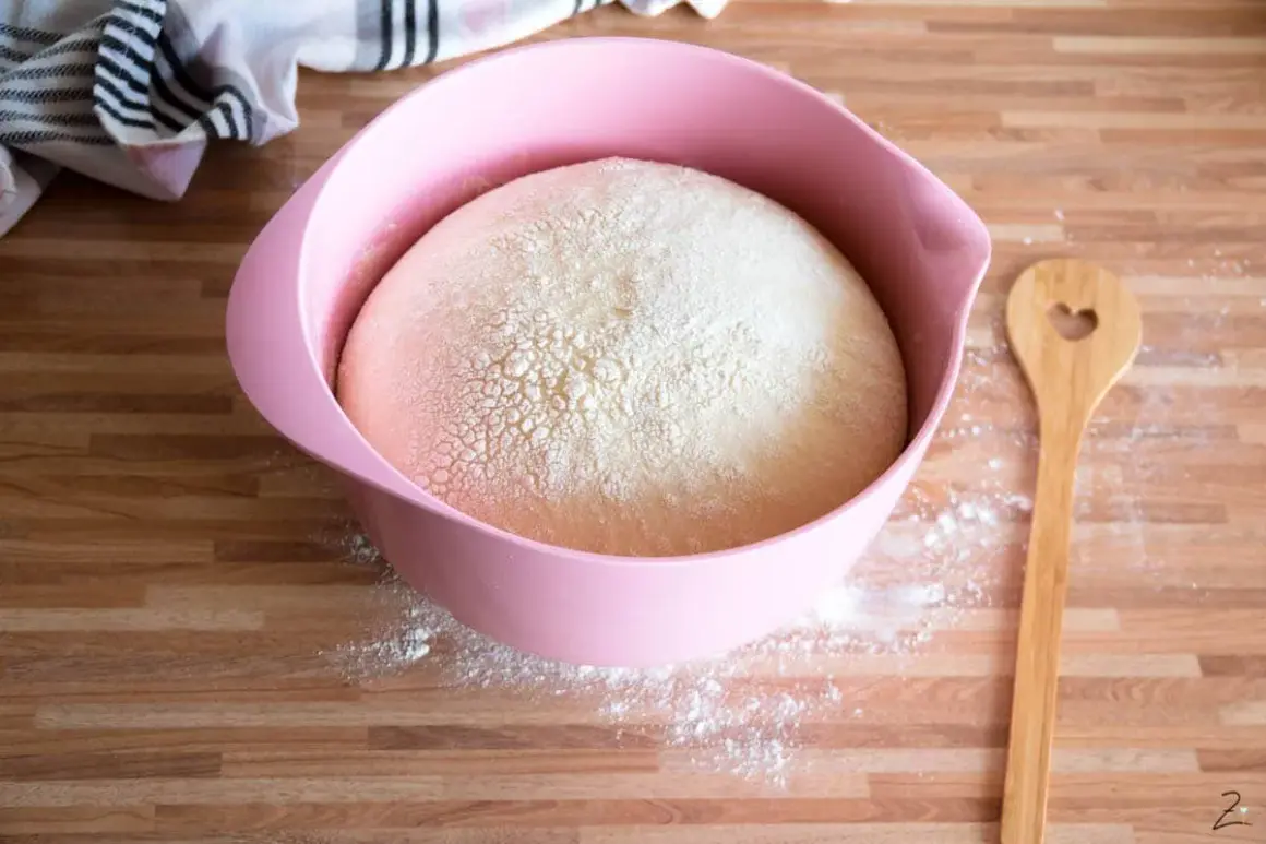 Ein aufgegangener Hefeteig mit Milch in einer rosa Schüssel, bereit zum Backen. Daneben liegt ein Holzlöffel mit Herz.