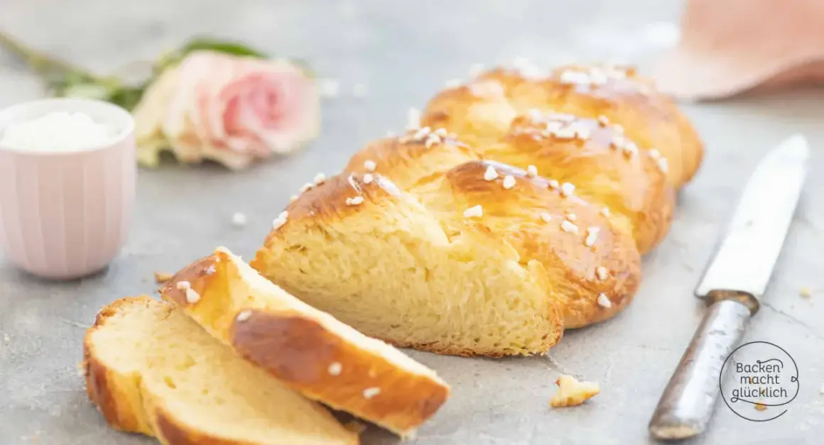 Ein goldbraunes Zopf Brot, bestreut mit Hagelzucker, liegt auf einem Tisch. Daneben eine rosa Rose und eine Tasse.