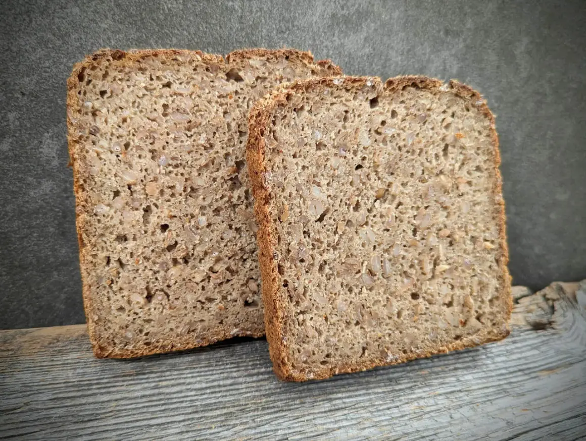 Zwei Scheiben rustikales **Kornbrot** liegen auf Holz. Das Brot ist dicht und voller Körner, mit einer dunklen Kruste.