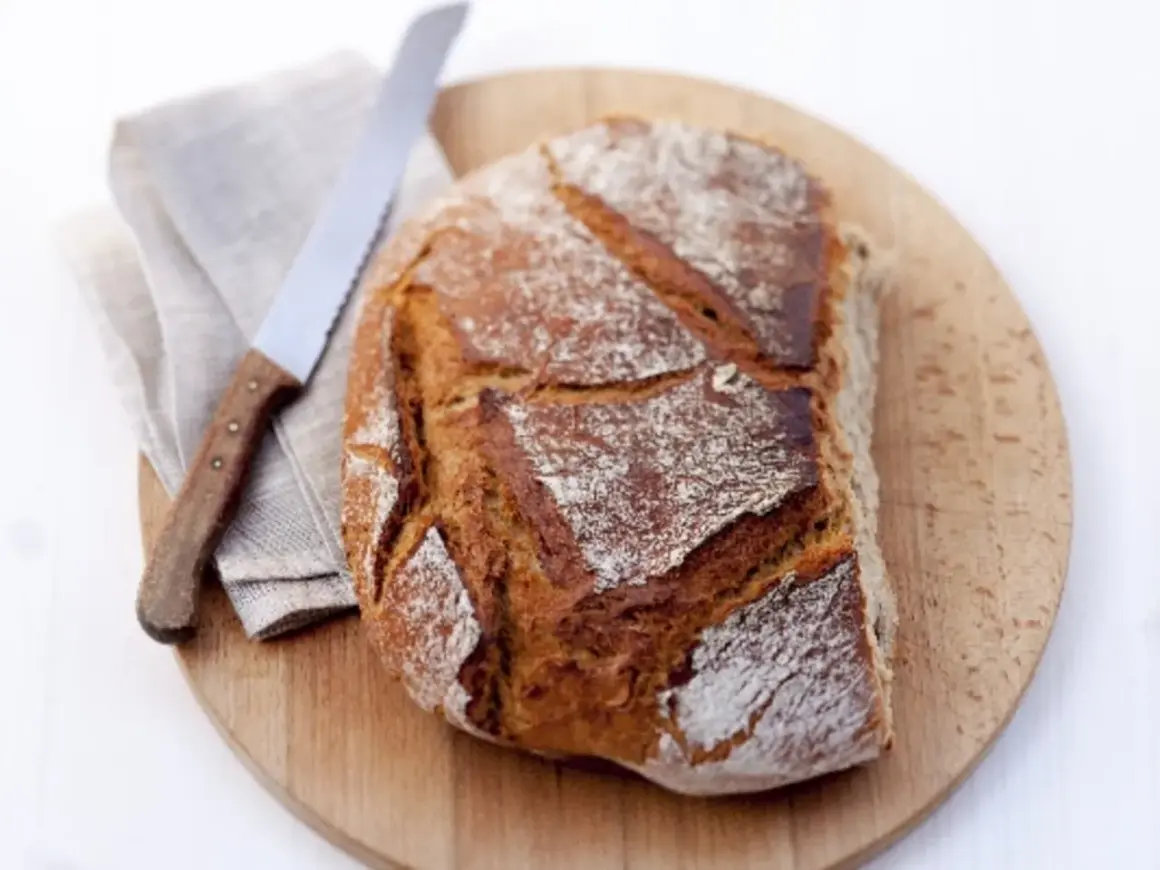 Ein frisch gebackenes Brot aus Hefe Teig liegt auf einem Holzbrett. Daneben ein Messer und eine Serviette.