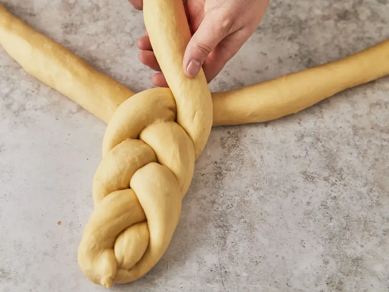 Eine Hand flechtet einen Osterkranz aus Hefeteig. Der goldgelbe Teig wird zu einem kunstvollen Zopf geformt.