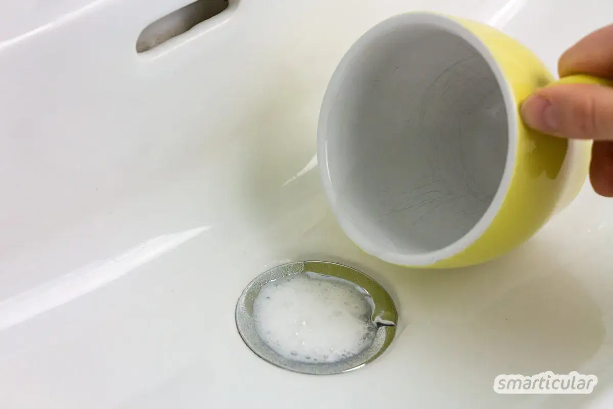 Gelbe Tasse kippt über einem Waschbeckenabfluss, Schaum bildet sich. Ein Hausmittel wie Essig oder Essigessenz hilft hier.