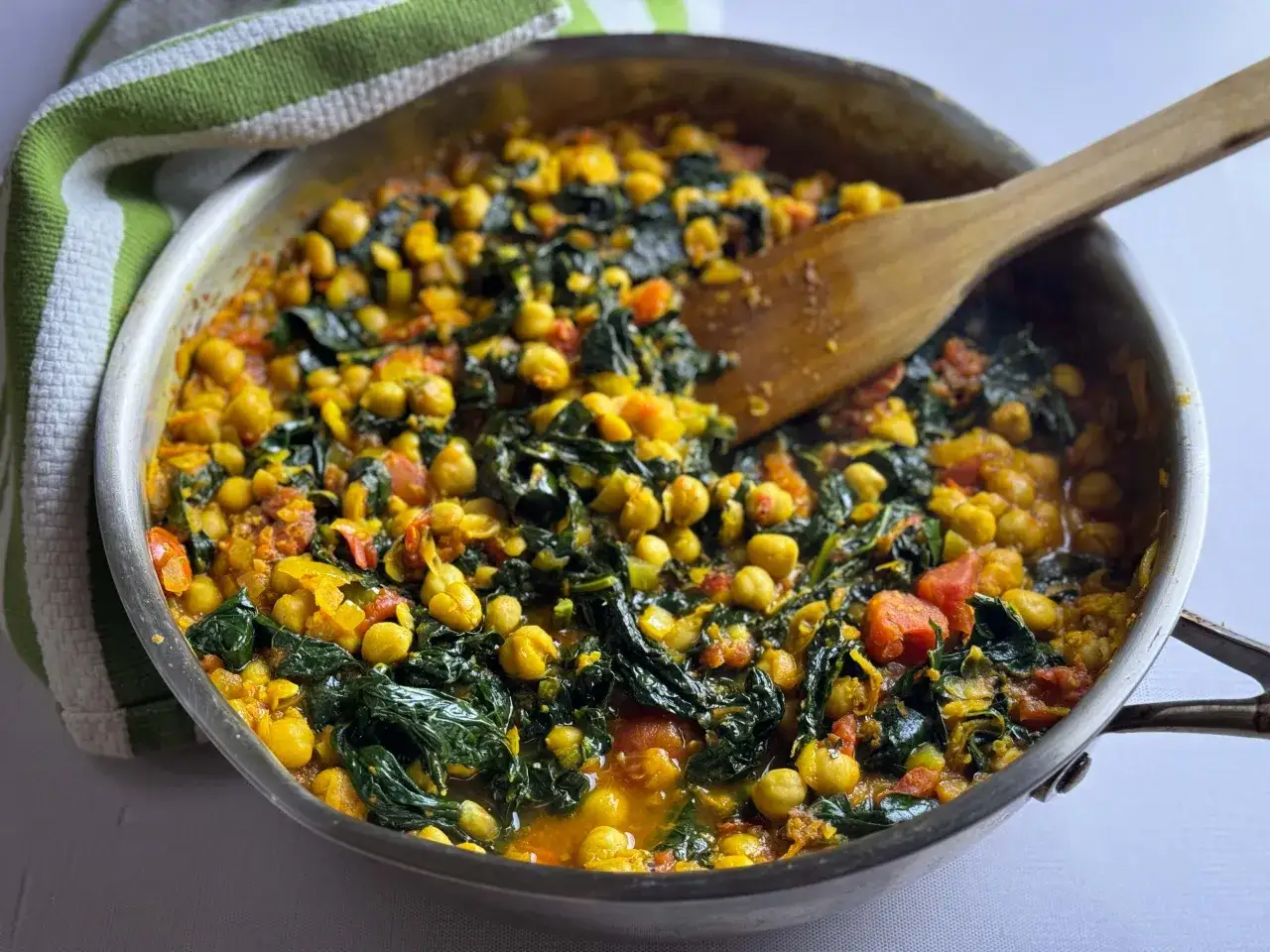 Herzhafte Kichererbsen mit Grünkohl und Tomaten in einer würzigen Sauce, inspiriert von Kurkuma-Wurzel-Rezepten. Ein Holzlöffel liegt in der Pfanne.