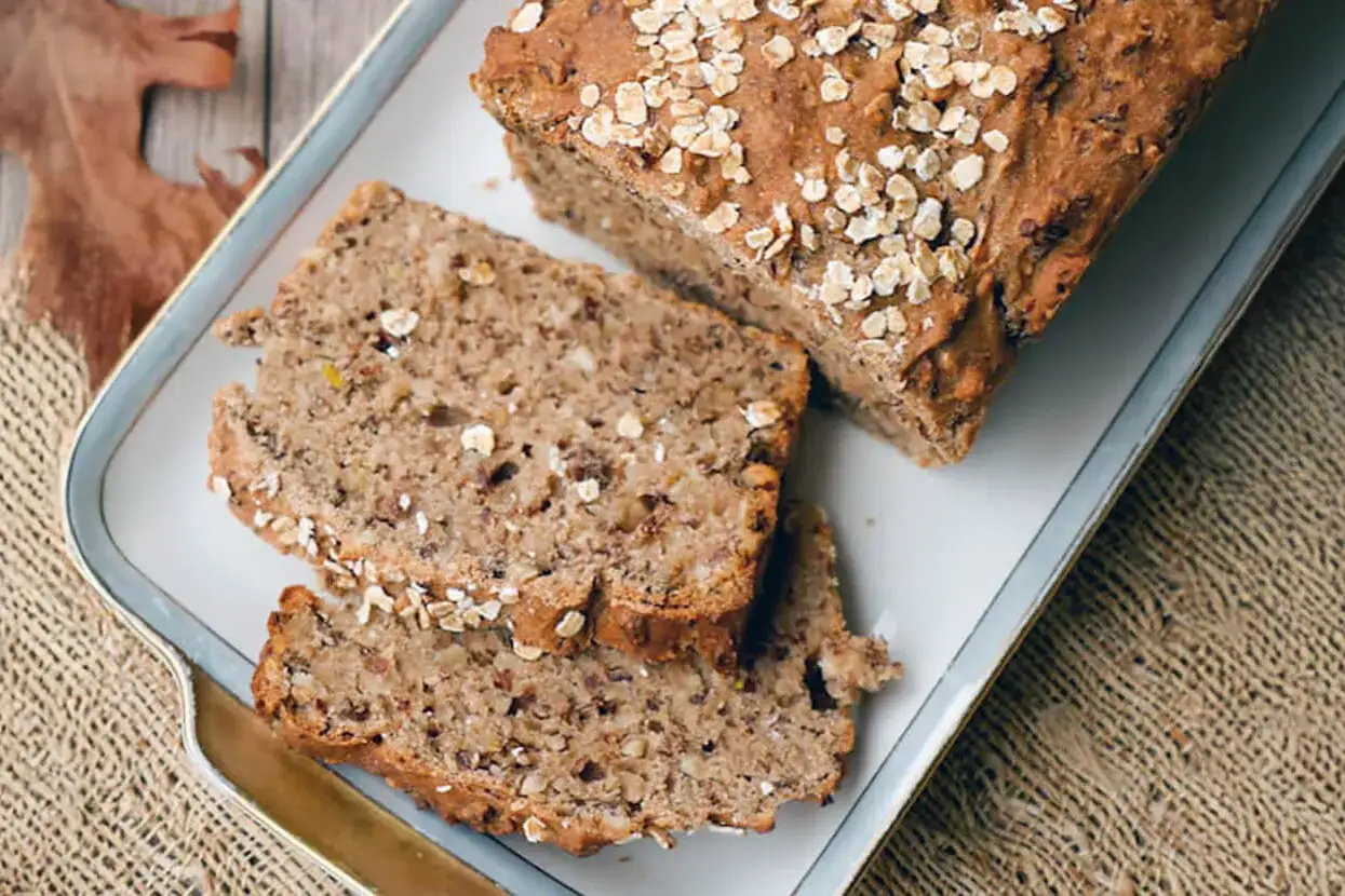 Ein rustikales Kornbrot mit Haferflocken und Nüssen, aufgeschnitten auf einem Teller, daneben ein Herbstblatt.