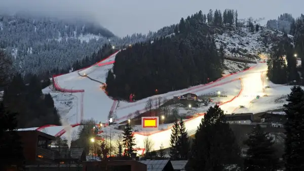 Die Skirennstrecke, beleuchtet für die Nacht, mit roten Banden und einem roten Hahnkamm-Symbol.