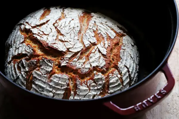 Frisch gebackenes Dinkel Mischbrot mit knuspriger Kruste und weißer Mehlschicht in einem roten Gusseisentopf.