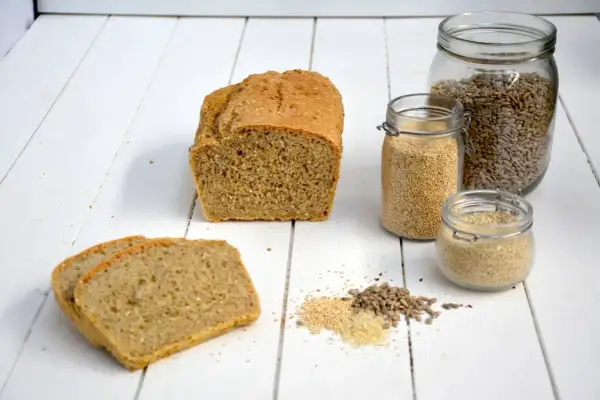 Brotlaib und Scheiben, daneben Gläser mit Quinoa, Sonnenblumenkernen und Sesam. Alles Zutaten für ein gesundes glutenfreies Mehl.